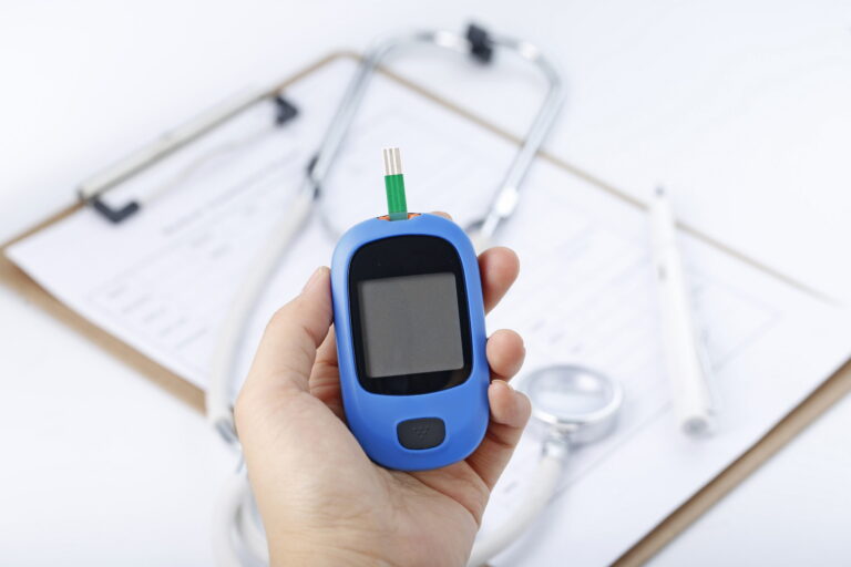 Hand holding a blood glucose meter measuring blood sugar, the background is a stethoscope and chart file