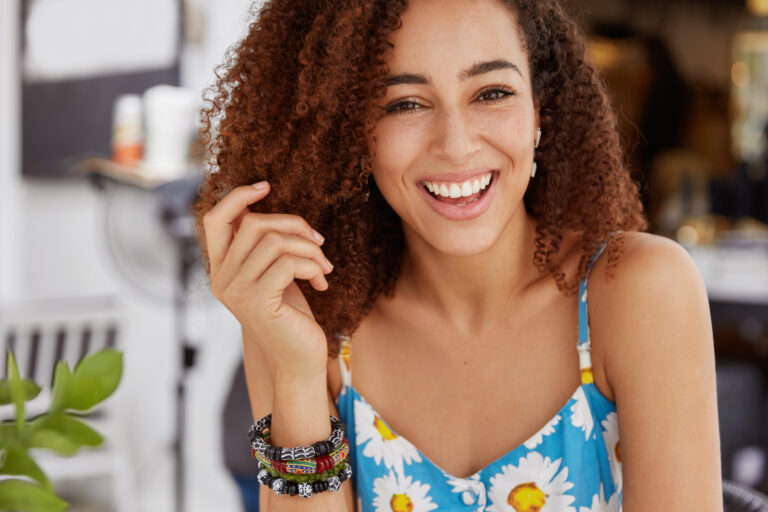 Joyful smiling dark skinned female with bushy hairdo, wears summer t shirt and bracelet, happy to spend free time in family circle or with friends, sits against cafe interior